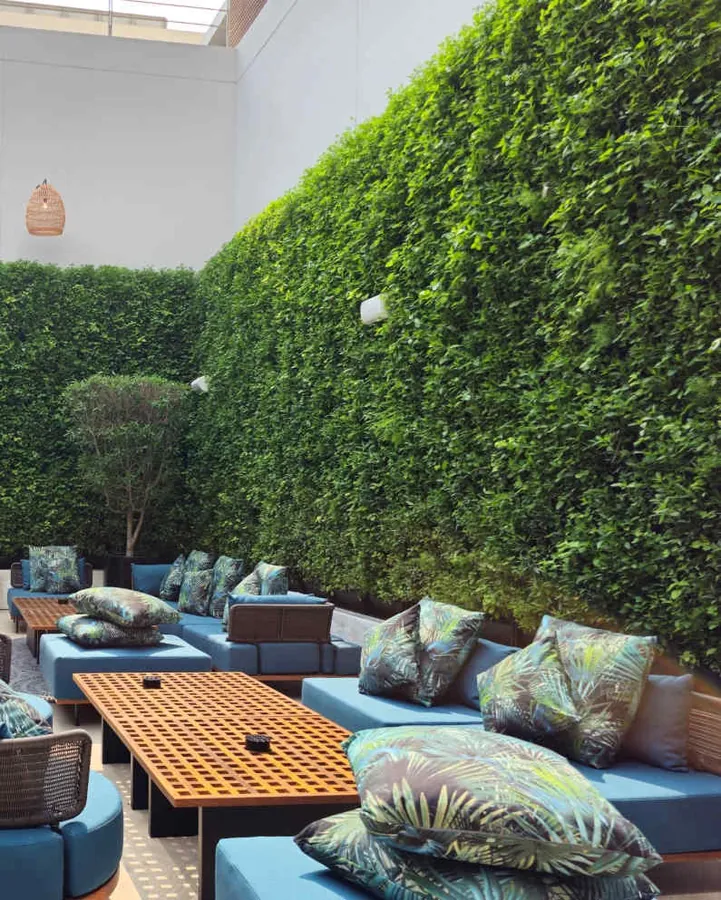 Tall green walls enclosing an open-air hotel lounge with blue sofas and timber tables